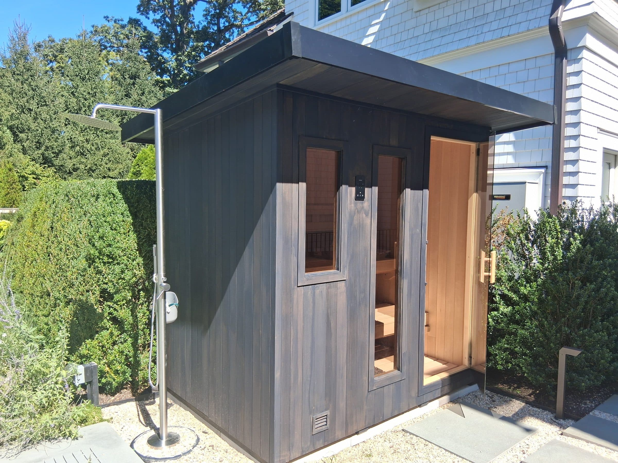 Dark outdoor sauna with an open glass door, outdoor shower, and stepping-stone approach.