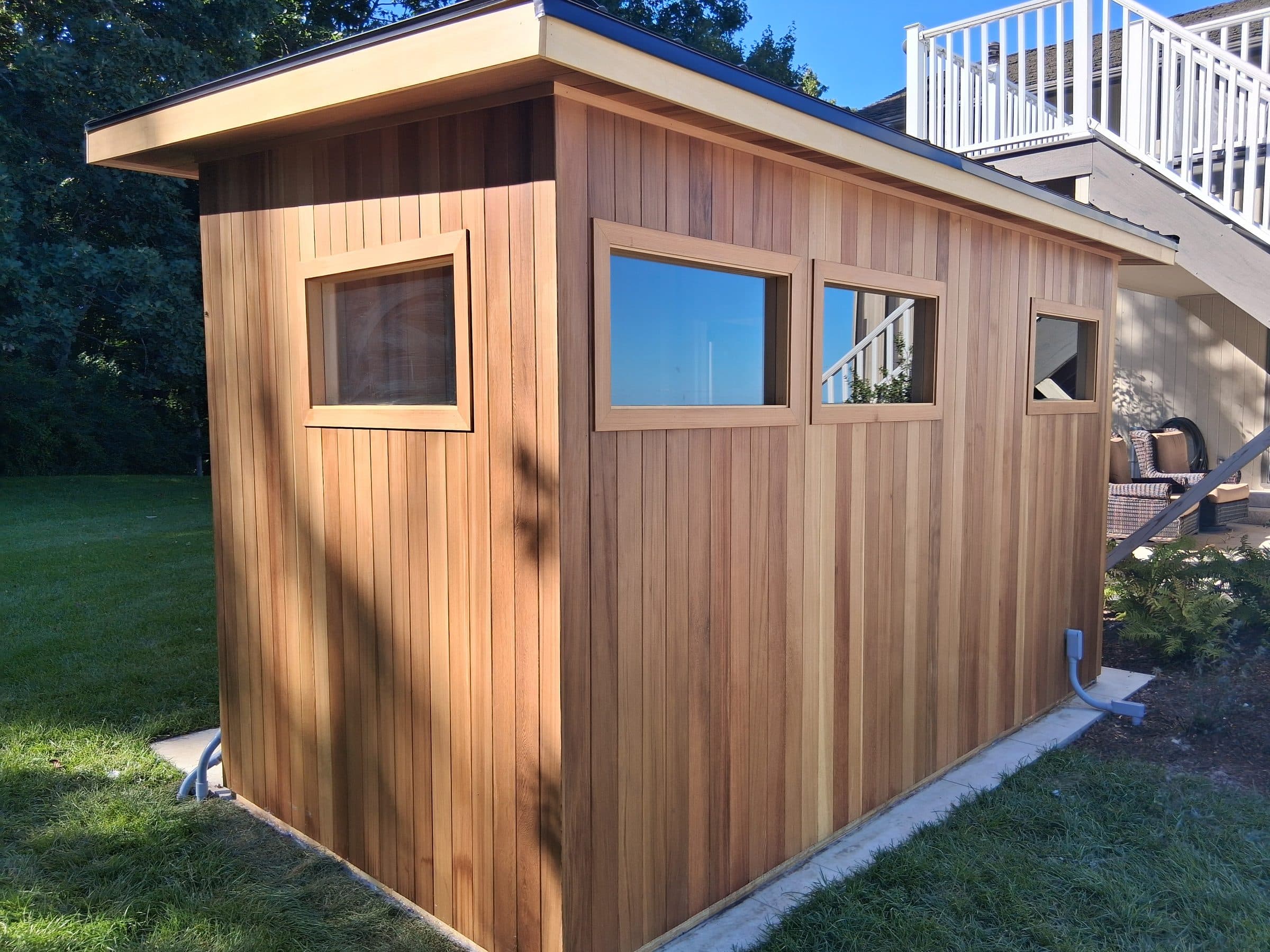 Natural wood outdoor sauna with several windows and a low roof on a prepared base beside a lawn.