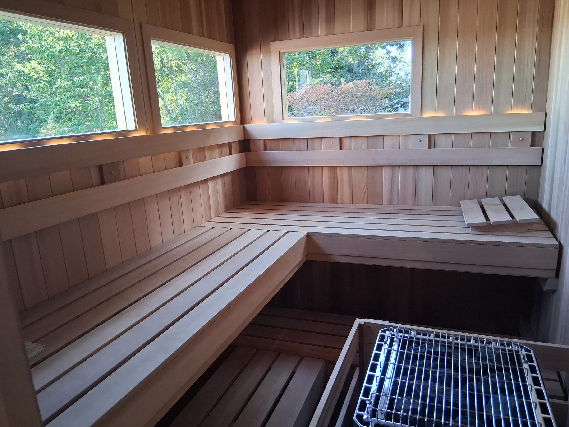 Sauna interior with L-shaped wood benches, three windows, backrest lighting, and a heater in the foreground.